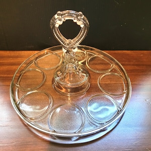 May include: A clear glass egg holder with a handle. The holder has six individual compartments for eggs and a central pedestal.