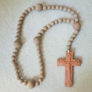 May include: A rosary featuring a series of matte, light tan spherical beads strung together, interspersed with larger beads of the same color. Attached to the end is a terracotta-colored cross with a raised relief figure of a person centered.