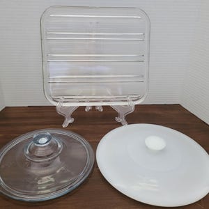 May include: Clear glass rectangular serving tray with horizontal ridges, displayed on a clear plastic stand. Two glass lids are also present: one clear with a knob, and one white with a white knob.