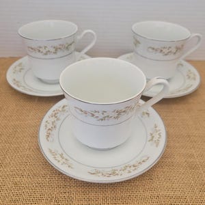 May include: A set of three ceramic mugs with a speckled, matte finish. The mugs are in shades of cream, light grey, and dark grey. Each mug has a simple, modern design and a rounded handle. The mugs are likely for coffee or tea.