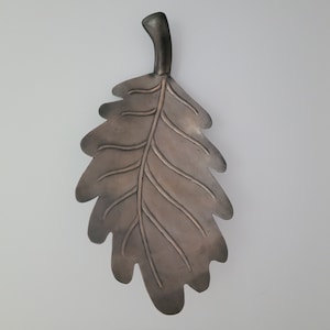 May include: A metal oak leaf wall decor with a textured surface and a raised stem. The leaf is a dark brown color.