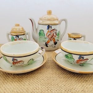 May include: A vintage porcelain tea set with a whimsical design featuring a boy carrying a pig. The set includes a teapot, creamer, sugar bowl, and two teacups and saucers. The teacups and saucers have a gold trim.