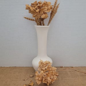Puede incluir: Un jarrón de cerámica blanco con base bulbosa y cuello delgado, con hortensias secas y espigas de trigo. El arreglo está sobre una superficie de arpillera, con pétalos caídos esparcidos.
