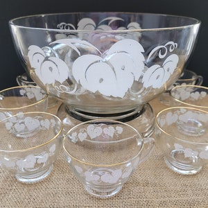 Może przedstawiać: Zestaw vintage szklanych naczyń do ponczu z wzorem białych winorośli. Miska ma złotą obwódkę i pasującą podstawę. Zestaw zawiera osiem pasujących filiżanek ze złotymi obwódkami.