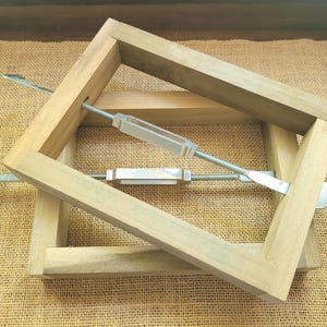 May include: Two rectangular wooden frames constructed from light-colored natural wood, each featuring a central metal turnbuckle mechanism with threaded rods and hook ends, designed for tensioning or securing the wooden frame components together.