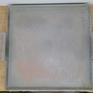 Puede incluir: Una plancha de metal cuadrada de color plateado con dos asas rectangulares. La plancha tiene una superficie ligeramente texturizada y parece ser utilizada para cocinar. Las asas tienen texto.