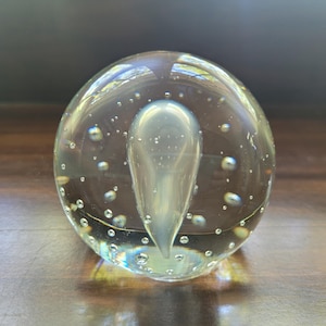 May include: A clear glass paperweight in a spherical shape featuring a central opaque white teardrop-shaped inclusion surrounded by numerous small air bubbles suspended throughout the transparent glass body, creating a layered internal visual effect.