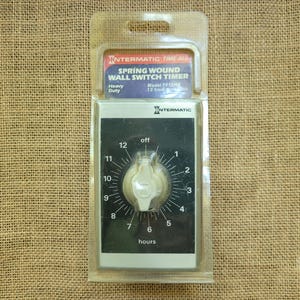 May include: An Intermatic spring wound wall switch timer in clear plastic packaging. The device features a black rectangular face with a white central dial, numbers 1 through 12, and the word hours printed below. The label reads Intermatic Time-All.