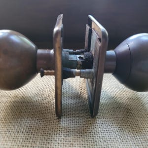 May include: Antique bronze door knob set with two rounded knobs and square backplates. The hardware shows signs of age and wear, with a visible screw and connecting mechanism. The set is likely for a vintage door.