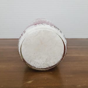 May include: A clear glass bottle with a white, textured interior. The bottle has a rounded base and a red-tinted rim. The words "CONTENTS" are visible on the side of the bottle. The bottle is resting on a wooden surface.