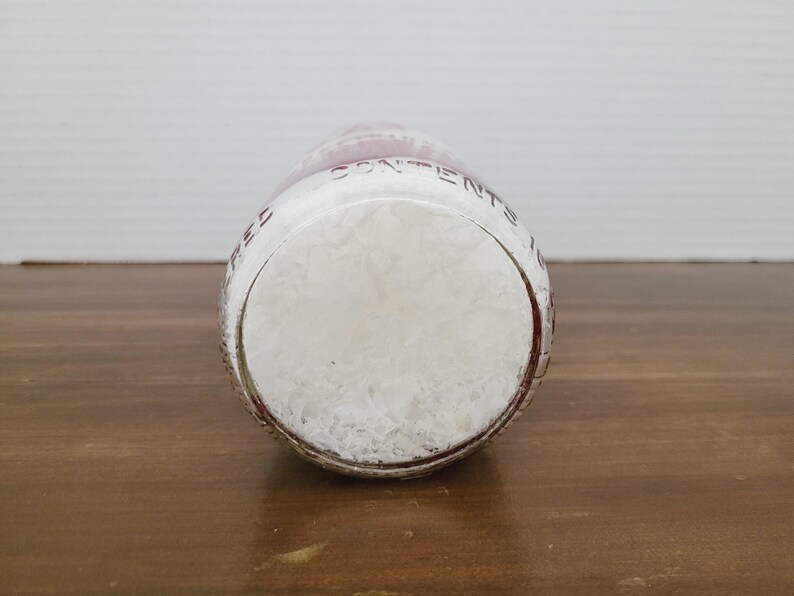 May include: A clear glass bottle with a white, textured interior. The bottle has a rounded base and a red-tinted rim. The words "CONTENTS" are visible on the side of the bottle. The bottle is resting on a wooden surface.