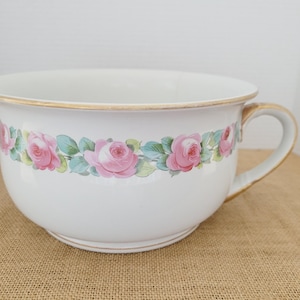 Op de afbeelding: Witte keramische kom met een gouden rand en handvat, versierd met een bloemenrozenpatroon in roze en groen. De kom heeft een klassiek ontwerp, geschikt voor decoratief of functioneel gebruik.