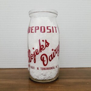 May include: Clear glass milk bottle with red lettering that reads "DEPOSIT" and "Rojek's Dairy". The bottle is filled with a white substance and has the address "5681 N. Tonawanda, N.Y." printed on it. The bottle is sitting on a wooden surface.