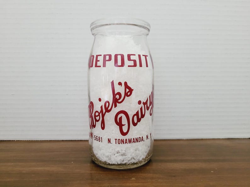 May include: Clear glass milk bottle with red lettering that reads "DEPOSIT" and "Rojek's Dairy". The bottle is filled with a white substance and has the address "5681 N. Tonawanda, N.Y." printed on it. The bottle is sitting on a wooden surface.