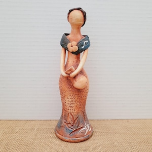 Puede incluir: Figura de cerámica de una mujer con un vestido marrón rojizo texturizado y un chal azul. La figura tiene un detalle floral en el chal y sostiene un objeto pequeño. La base presenta un diseño floral con detalles blancos.