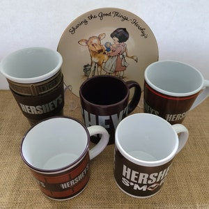 May include: Five ceramic mugs with Hershey's branding. The mugs are brown and white with different designs. One mug has a Hershey's S'mores design. Another mug has a Hershey's chocolate bar design. The mugs are on a brown burlap surface.  The text on the plate reads "Sharing the Good Things - Hershey's".