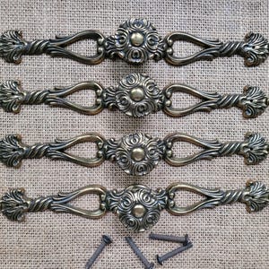 May include: Four antique bronze drawer pulls with ornate floral and scrollwork designs. Each pull features a central circular medallion and curved handles. Includes mounting screws.