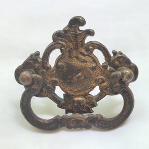 May include: A dark bronze-toned metal drawer pull featuring an ornate, symmetrical backplate with intricate scrollwork and floral detailing. The hardware includes a hinged, curved drop handle attached to two circular mounting points on the base.