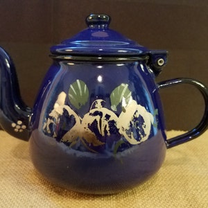 Puede incluir: Una tetera de esmalte azul con un diseño floral blanco. La tetera tiene una tapa y un asa.