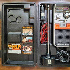 May include: A General Electric 3-5000 emergency radio kit in a black case. The kit includes a radio with an orange and silver face, a car adapter, and an antenna. The case has a "HELP!" sticker and instructions.
