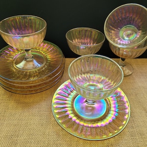 May include: A collection of iridescent glass dessert dishes and plates. The set includes stemmed bowls and matching plates, all with a ribbed design. The glass reflects a rainbow of colors, creating a shimmering effect. The dishes are arranged on a textured surface.