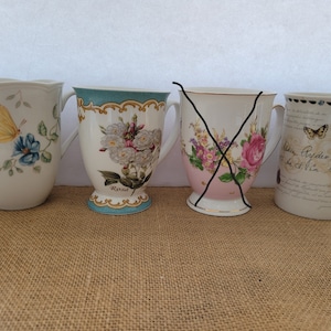 May include: Four decorative ceramic mugs with floral and butterfly designs. One mug features a yellow butterfly and blue flowers. Another has white roses and the word "Rosa". The third mug has a butterfly and script. The fourth mug is crossed out.