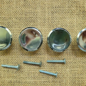 May include: Four circular cabinet knobs with a polished silver-tone metallic finish and a smooth, reflective surface. Each knob is accompanied by a matching silver-tone metal screw, positioned directly below the corresponding knob hardware.