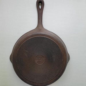 Könnte beinhalten: Eine braune gusseiserne Bratpfanne mit langem Griff und kreisförmiger Kochfläche. Der Boden der Pfanne hat ein strukturiertes Muster und die Worte "Genuine Cuisine" sind eingeprägt. Der Griff hat ein Loch zum Aufhängen.