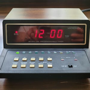 May include: A black and brown Micronta Seven Day Programmable Timer with a digital display showing 12:00 in red. The timer has several buttons and indicator lights for various functions, including time setting and program selection.
