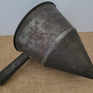 Peut inclure: Une ancienne entonnoir en métal avec une poignée. L'entonnoir a un large sommet rond qui se rétrécit en un tamis conique en forme de cône. Le métal présente un aspect vieilli, avec des zones de rouille et de décoloration. La poignée est cylindrique et fixée sur le côté.