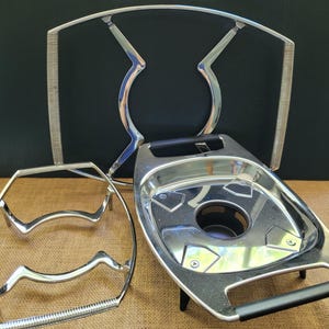 Könnte beinhalten: Drei silberne Metallteile für eine Kaffeemaschine. Das größte Teil ist eine Tropfschale mit einem Griff. Das mittlere Teil ist ein runder Metallring. Das kleinste Teil ist ein Metallrahmen mit zwei gebogenen Armen.