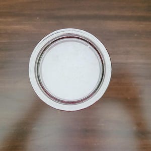 May include: Overhead view of a clear glass jar lid with a white interior. The lid has a silver rim and a red accent ring. The jar lid is centered on a brown wooden surface.