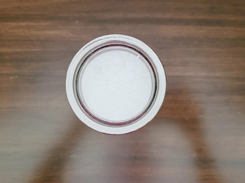 May include: Overhead view of a clear glass jar lid with a white interior. The lid has a silver rim and a red accent ring. The jar lid is centered on a brown wooden surface.
