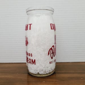 May include: A clear glass milk bottle filled with white material. The bottle has red lettering that reads "Delicious Cream" and "Reid's". The bottle is cylindrical with a rounded top and is sitting on a wooden surface.