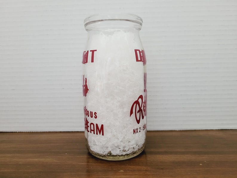 May include: A clear glass milk bottle filled with white material. The bottle has red lettering that reads "Delicious Cream" and "Reid's". The bottle is cylindrical with a rounded top and is sitting on a wooden surface.