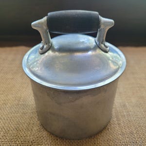 May include: A cylindrical aluminum container with a smooth, metallic finish and a matching lid. The lid features a central handle with a dark, cylindrical grip secured by two silver-toned metal brackets attached to the top of the lid surface.