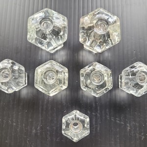 May include: A collection of eight clear, faceted glass cabinet knobs. Each knob has a hexagonal shape with a central hole for mounting. The knobs are arranged on a dark surface, showcasing their reflective qualities.