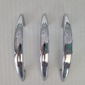 May include: Three polished chrome cabinet pulls arranged vertically. Each handle features a smooth, arched, crescent-like shape with a reflective metallic finish. The design is simple and curved, showing a clear, mirror-like surface that reflects the surroundings.