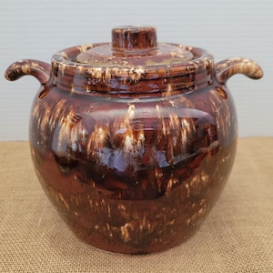 Peut inclure: Un grand pot en céramique marron avec un couvercle et deux poignées. Le pot a une finition brillante avec un effet de glaçure dégoulinante dans les tons de marron et de blanc. Le couvercle a un petit bouton pour faciliter le retrait.