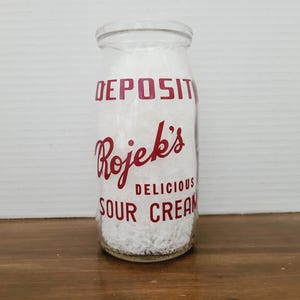 May include: A clear glass milk bottle filled with white contents, likely sour cream. The bottle has red lettering that reads "DEPOSIT" and "Rojek's DELICIOUS SOUR CREAM". The bottle is sitting on a wooden surface.