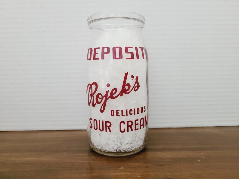 May include: A clear glass milk bottle filled with white contents, likely sour cream. The bottle has red lettering that reads "DEPOSIT" and "Rojek's DELICIOUS SOUR CREAM". The bottle is sitting on a wooden surface.