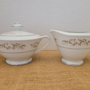 May include: A white porcelain sugar bowl with a lid and a creamer. Both pieces feature a floral design in brown and gold, with a silver trim. The sugar bowl has two handles and a lid with a small knob.
