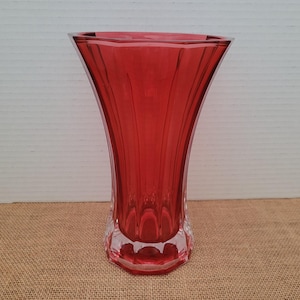 May include: A tall, ruby red glass vase with a flared top and a clear, faceted base. The vase has vertical ridges that run from the top to the base. The vase is approximately 10 inches tall.