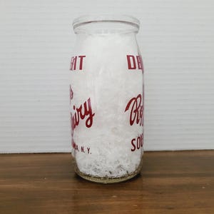May include: A clear glass milk bottle with red lettering. The bottle is filled with a white substance. The text on the bottle includes the words "Dairy" and "Souda, N.Y."