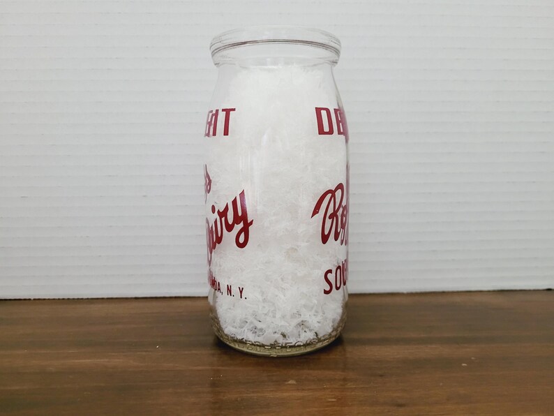 May include: A clear glass milk bottle with red lettering. The bottle is filled with a white substance. The text on the bottle includes the words "Dairy" and "Souda, N.Y."