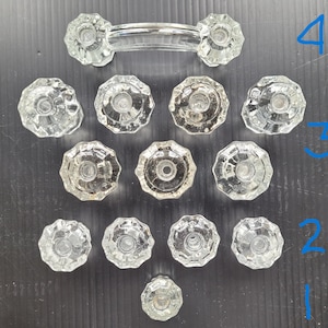 May include: A collection of clear glass cabinet knobs and a matching handle. The knobs have a faceted, flower-like design. The handle is a curved bar with knobs on each end. The hardware is arranged on a dark surface.