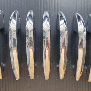 May include: Seven chrome-plated door handles arranged on a black surface. The handles have a curved design with a shiny, reflective surface. The handles are likely for a vehicle or furniture.