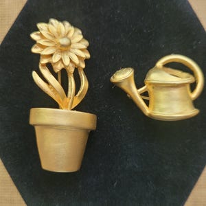 May include: Two gold-toned metal brooches. One is a flower in a pot, the other is a watering can. The flower brooch features a detailed daisy-like bloom atop a stem with leaves, set in a simple pot. The watering can brooch has a handle and spout.