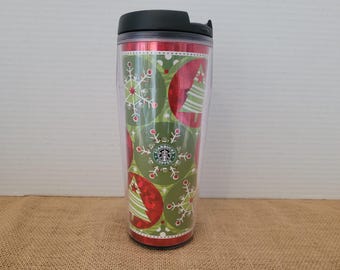 May include: A red and green reusable travel mug with a Christmas tree and snowflake design. The mug features the Starbucks logo and the text "Starbucks Coffee".