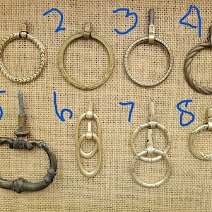 May include: A collection of eight metal drawer pulls in various brass and bronze finishes, each featuring a ring or oval handle attached to a mounting post. The pulls are numbered one through eight in blue ink, showcasing diverse textures and shapes.